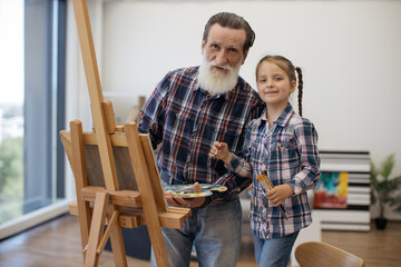 Portrait of happy aged grandpa holding palette with mixed colors and smiling girl with brushes in home interior. Experienced senior painter rejoicing in creativity with granddaughter in living room.