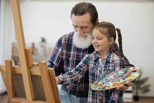 Cheerful older adult with palette hugging small tween with brush making strokes on cloth at easel. Delighted grandfather teaching girl basics of painting while building new experiences together.