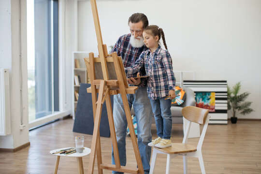 Full length view of grey-haired man preparing paint on palette while stylish girl standing in his hug. Relaxed child in denim outfit taking pleasure of art making with grandpa in studio at home.