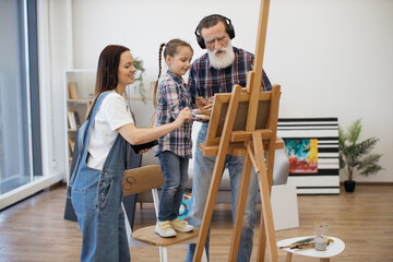 Modern pensioner in headphones and smiling lady with wired earbuds dancing with cute girl while...