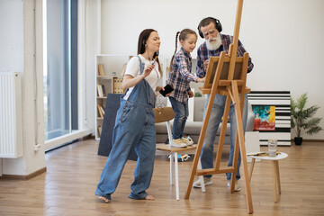 Obraz na płótnie Canvas Excited senior man and young females with devices and paintbrushes moving to beat of songs behind easel. Thrilled relatives getting inspired by tunes before starting new art project in home interior.