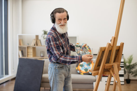 Cheerful elderly man in jeans using brush and palette while painting with audio accompaniment in headphones. Grey-haired professional expressing mastery through colors on canvas in art space.