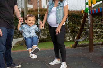 Happy joyful smiling little toddler playing on a swing with both her parents and her pregnant mother expecting her future sibling.