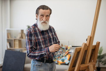 Portrait of confident bearded pensioner with brush posing for camera during creative process in studio. Efficient painter playing freely with ideas, marks, colors and surfaces for success of artwork.