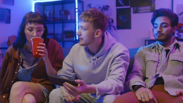 Medium shot of young Caucasian girl and two diverse male friends sitting together on couch with smartphone, watching sport game, cheering anxiously, then getting disappointed at missed goal