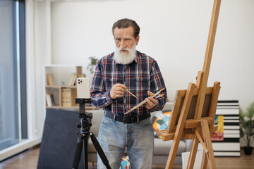 Creative pensioner blogging own painting technique and skill while staying in comfortable home studio. Senior artist looking at camera of cellphone attached to black tripod and recording video guide.