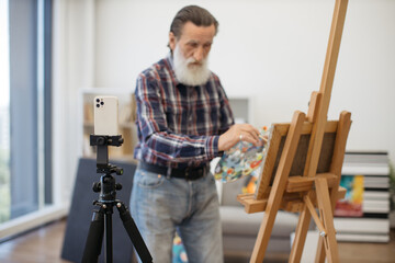 Elderly male painter with long grey beard using mobile phone with tripod for shooting video for family archive. Senior man dressed in jeans and checkered shirt demonstrating drawing skills on camera.