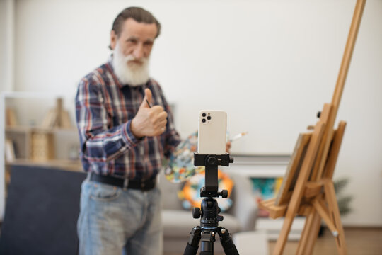 Charismatic retired man wearing checkered shirt and jeans recording video while drawing in stylish art studio. Grey-bearded artist holding palette with paintings and showing thumb up.