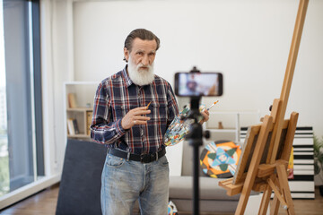 Aged gentleman with long grey beard standing in front of tripod with mobile phone and telling about drawing techniques in art. Casually dressed professor with palette in hands holding online courses.