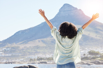 Woman at beach, arms in air and freedom, back view with mountains and travel, praise and sunshine. Female person outdoor, carefree and adventure, mindfulness and peace with worship in nature