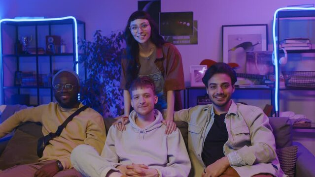 Medium portrait shot of three multiethnic diverse young friends sitting together on couch in student dorm, in blue neon light, Caucasian girl walking up and hugging man, smiling and looking at camera