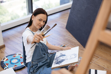 Portrait of charming female painter in denim jumpsuit and white t-shirt sitting on floor and...
