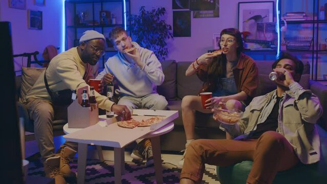 Full shot of four young multiethnic men and woman in casual clothes sitting together on couch and chairs at student home party in blue neon light, eating pizza, drinking beer and chatting