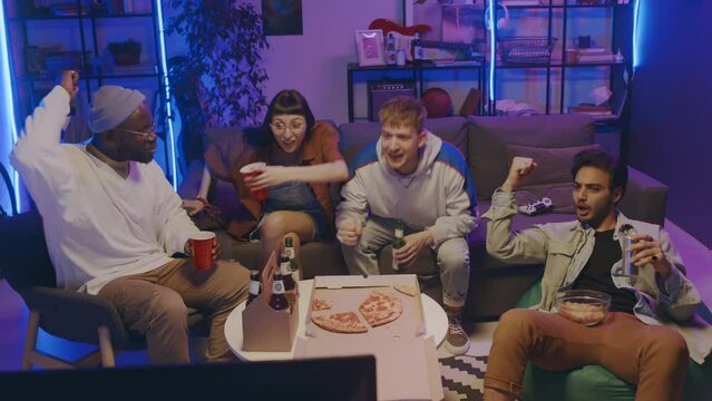 Full shot of young multiracial students sitting together at home party, eating pizza, drinking beer, watching sport match on TV, cheering, then shouting with joy and hugging at winning goal