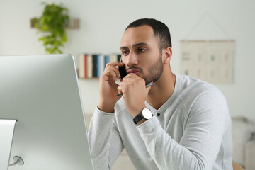 Young man talking on smartphone while working with computer in room. Home office