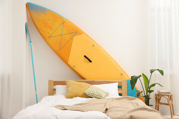 Large comfortable bed, SUP board and green houseplant in stylish bedroom. Interior design