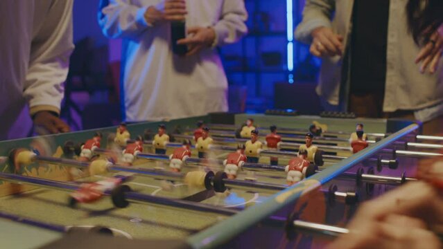 Close-up shot of unrecognizable young multiracial girl and guy playing table football, and anonymous friends watching and cheering
