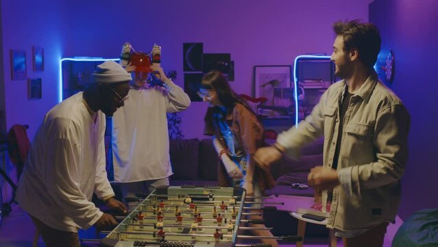 Two ethnically diverse young men playing table football at student party in blue neon light, Arab guy winning, Caucasian couple with fun beer hat watching and cheering