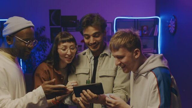 Medium close-up shot of group of ethnically diverse young people in casual clothes standing close together and watching sports game on smartphone, cheering, whooping and toasting with beer at goal