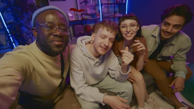 Medium shot of four happy young Caucasian, African American and Arab friends hanging out together in student dorm room, recording story for social network, smiling and talking