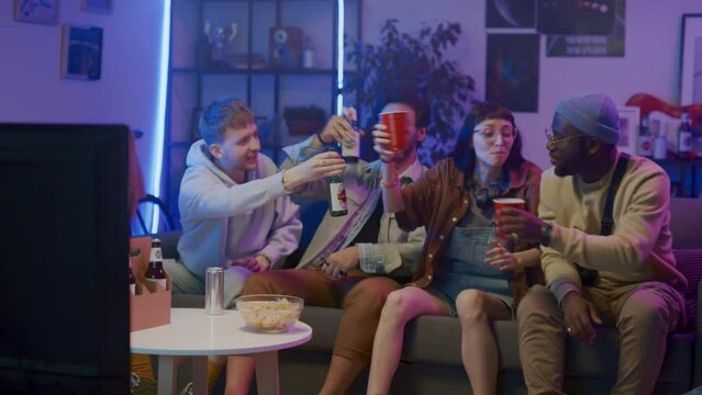 Full shot of four multiracial friends sitting on couch in living room with purple neon light, watching football match on TV, cheering, shouting loudly and toasting with beer at winning goal