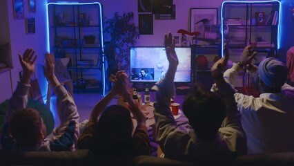 Full rear shot of four diverse young guys and girl sitting on couch in dark room with blue neon light, watching cybersport championship stream of FPS shooter on TV, cheering and clapping