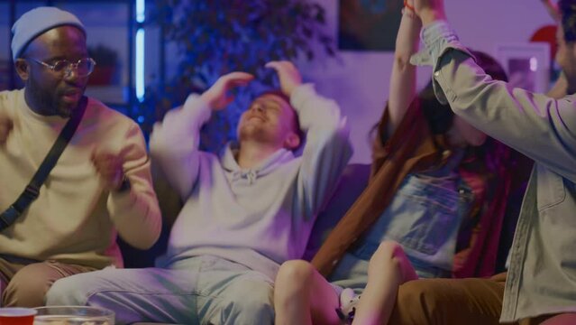 Medium shot of young multiethnic friends sitting on couch in front of TV, playing fighting video game on console with wireless controllers, cheering, Caucasian girl winning and hugging boyfriend