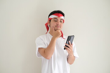 A young Asian man wearing a white T-shirt and Kemerdekaan Indonesia costume is thinking while looking at his smartphone. Indonesia Independence Day. Isolated white background.