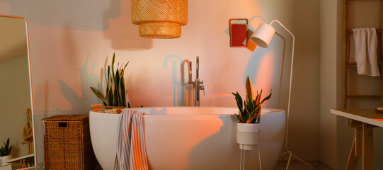 Interior of modern bathroom with bathtub, lamps and houseplants in evening