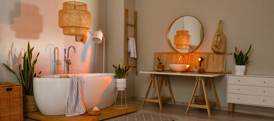 Interior of light bathroom with bathtub, table, sink and houseplants in evening