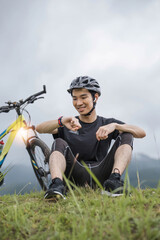 Fototapeta premium happy young Asian man in sportswear checks his calories burned on his smartwatch while resting on the grass after a long bike ride in the mountains.