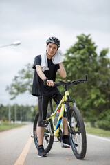A tired and sweaty young Asian man in sportswear and a bike helmet wiping his neck with a towel while biking along the country roads.