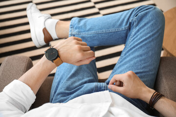 Stylish young man sitting on armchair looking at wristwatch in room