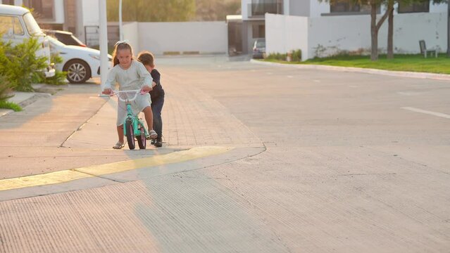 par de ni&ntilde;os hermanos amigos primos latinos mexicanos jugando y ayudando a andar aprender en bicicleta por los suburbios de la ciudad en la calle en vacaciones en el verano al aire libre al atardecer