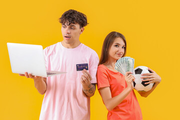 Young couple with soccer ball, laptop and money on yellow background. Sports bet concept