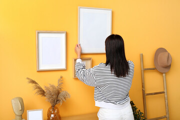 Young woman hanging blank frame on yellow wall in living room, back view