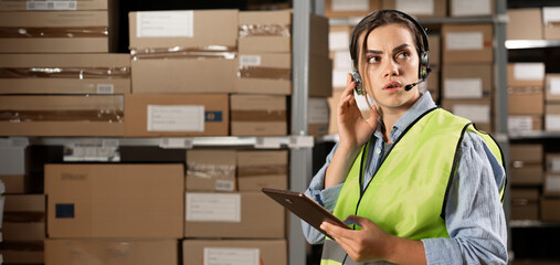 brazilian female worker in headset at logistics warehouse using digital tablet. speaks into a microphone against the background of shelves with cardboard boxes.