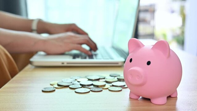 A pink piggy bank on table with someone working on laptop in the background. 