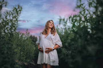 Pregnant woman in white boho summer dress walks in the middle of the field in the tall green grass
