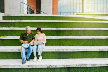 Daughter and father sit together in the vacant space on campus spend their free time teaching senior father to learn how to read health care for old age father understand simple use of social media.