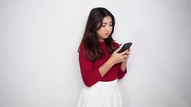 A thoughtful young Asian woman is wearing red t-shirt and holding her phone looks confused between choices, isolated by white background