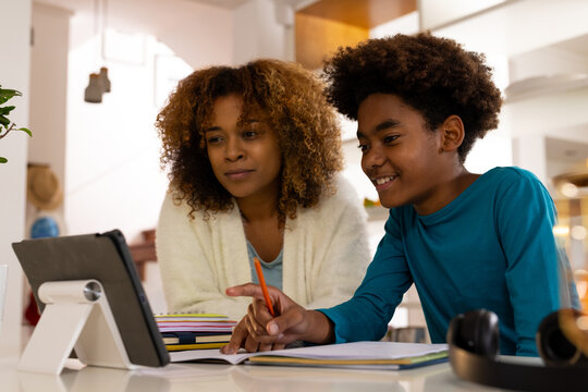 Happy african american mother and son sitting at table, using tablet, doing homework