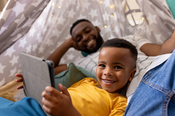 Happy african american father and son using tablet in tent at home