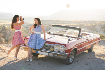 two beautiful girl in a retro dress are photographed on camera  on  mountain landscape in Cappadocia near a red retro car