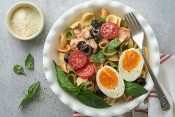 Italian pasta salad. Orecchiette pasta with tuna, tomato cherry, olive, basil and parmesan cheese in plate on grey stone or concrete background. Traditional Italian cuisine. Flat lay. Copy space.