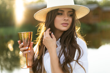 beautiful glamorous girl in a white dress and a straw hat on the shore of the lake with a goblet in...