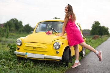 beautiful girl in a pink dress with a bouquet of red roses near a yellow retro car