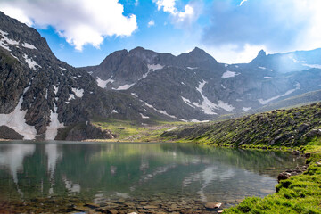 Mountain lake and rocky mountains. Crystal clear lake in the middle of the rocks. Green fields, lake and mountain ranges under snow