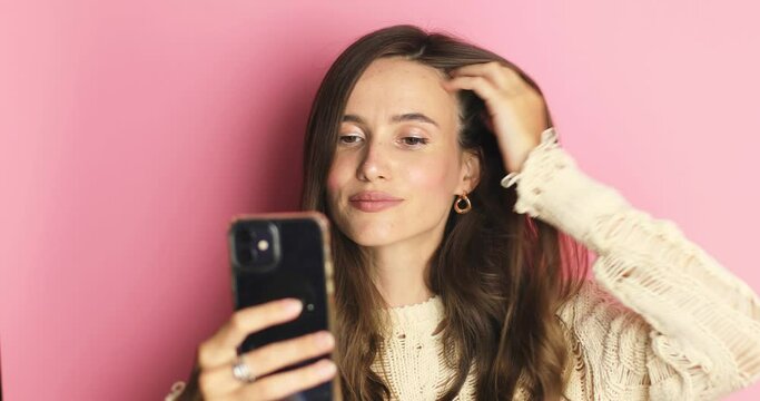 The girl stares at the phone and fixes her hair. Female uses phone like mirror. Young girl make photo from hands with phone on pink background, look at camera and smiling.