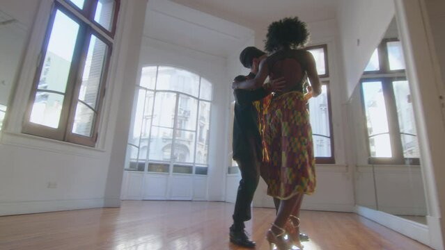 Young beautiful couple in stage outfits dancing Argentine tango in a spacious sunlit studio. Full length shot, lens flare